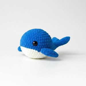 May include: A handmade blue and white crochet whale toy with black eyes. The plush whale has a rounded body and small fins. The toy is made of soft yarn and is suitable for children.