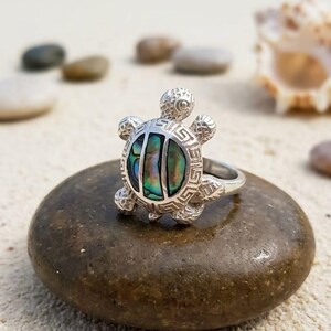 May include: A silver ring featuring a turtle-shaped centerpiece with an iridescent abalone shell inlay. The turtle's shell is divided into three sections, surrounded by a silver border with a geometric Greek key pattern and textured flippers.