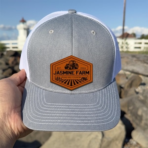 May include: A grey and white trucker cap with a leather patch. The patch displays the text "Jasmine Farm" with a tractor graphic and "Texas Hill Country" below. The cap has a curved peak and a mesh back.