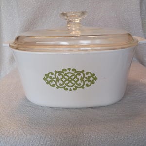 May include: White casserole dish with a clear glass lid and handle. The dish features a green decorative design on the front. The lid has a clear glass knob. The dish is likely made of ceramic or a similar material.