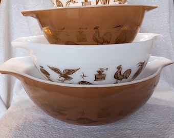 May include: A stack of four vintage Pyrex mixing bowls. Two white bowls and two brown bowls are nested together. The white bowls have a brown eagle, corn, and rooster design. The brown bowls have a similar design.