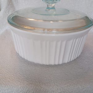 May include: A white, ribbed casserole dish with a clear glass lid and a small, round handle. The lid has a decorative pattern around the edge. The dish is designed for baking and serving.
