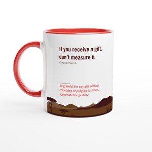 Peut inclure: Mug en céramique blanche avec intérieur et anse rouges. Le mug présente le texte "If you receive a gift, don't measure it" et un proverbe kenyan. Un paysage brun est au bas du mug.