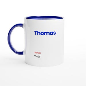 Pode incluir: Caneca de cerâmica branca com alça e borda azuis. A palavra "Thomas" está impressa em azul, com "Aramaic Twin" em vermelho e preto abaixo. A caneca é projetada para bebidas.