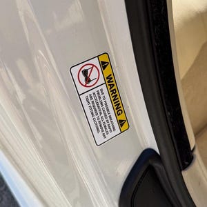 Peut inclure: Une étiquette d'avertissement rectangulaire avec un bord jaune et du texte noir est apposée sur une portière de voiture blanche. L'étiquette met en garde contre d'éventuels problèmes respiratoires dus à une accélération rapide et conseille de retirer les vêtements moulants.