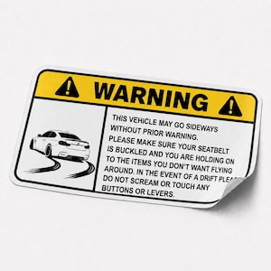 Op de afbeelding: Een rechthoekige sticker met een gele koptekst met het woord WARNING en twee uitroepteken-iconen. De tekst luidt: THIS VEHICLE MAY GO SIDEWAYS WITHOUT PRIOR WARNING. PLEASE MAKE SURE YOUR SEATBELT IS BUCKLED AND YOU ARE HOLDING ON.