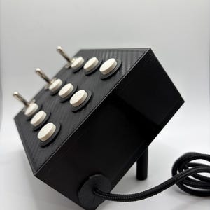 Könnte beinhalten: Ein schwarzes elektronisches Gerät mit strukturierter Oberfläche, das mehrere weiße Knöpfe und silberne Kippschalter aufweist. Ein schwarzes Kabel erstreckt sich von der Seite. Das Gerät ist abgewinkelt und zeigt sein Design und seine Komponenten.