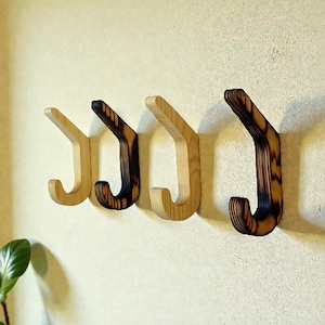Può includere: Quattro ganci da parete in legno in varie tonalità di marrone sono montati su una parete di colore chiaro. I ganci hanno un design curvo, con le venature del legno visibili. I ganci sono distanziati in modo uniforme, creando una soluzione di stoccaggio decorativa e funzionale.