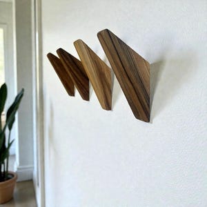 Puede incluir: Cuatro perchas de pared de madera en fila, cada una con forma triangular. Las perchas son de madera marrón claro con vetas visibles, montadas en una pared blanca. Las perchas están anguladas, creando un diseño moderno y minimalista.