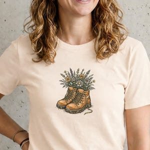 Puede incluir: Una camiseta color crema con un gráfico de botas de montaña marrones llenas de flores. Las botas tienen cordones y están detalladas con sombreado. Las flores son una mezcla de blanco, amarillo y morado.