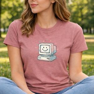 Può includere: T-shirt rosa antico con grafica di computer retrò con un volto sorridente. La grafica è blu, bianca e grigia. La persona indossa jeans blu.