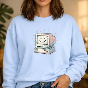 Könnte beinhalten: Hellblaues Sweatshirt mit einem Retro-Computer-Motiv. Der Computer zeigt ein lächelndes Gesicht auf dem Bildschirm, mit rosa, blauen und weißen Akzenten und funkelnden Sternendetails. Das Sweatshirt hat eine einfarbige hellblaue Farbe.