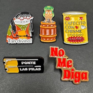 May include: A collection of colorful enamel pins featuring cartoon characters and phrases. The pins include a character with the text "Tan divino!", a character in a barrel, a yellow can with "Cafecito con Chisme", a black pin with "Ponte las Pilas", and a red pin with "No Me Diga".