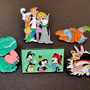 May include: Collection of enamel pins featuring cartoon characters. Includes a green dinosaur with a pink heart, a family from The Jetsons, characters from Animaniacs, Patrick Star, and Ren & Stimpy.