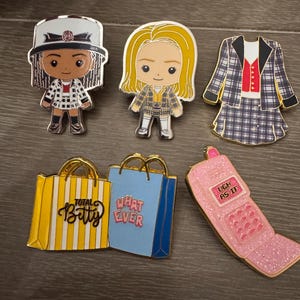 May include: A collection of enamel pins featuring characters and iconic items from a movie. Includes character pins, shopping bags with text "Total Betty" and "What Ever", and a pink flip phone pin with the text "Ugh As If".