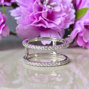 May include: A silver ring with two bands, each encrusted with small, clear gemstones. The ring is set against a backdrop of soft, purple flowers, creating a delicate and elegant aesthetic. The ring's design is modern and eye-catching.