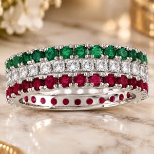 Puede incluir: Un anillo de eternidad de varias filas con tres hileras distintas de piedras preciosas de corte redondo engastadas en metal plateado. La fila superior tiene piedras verdes, la fila central piedras blancas y la fila inferior piedras de color rojo intenso.