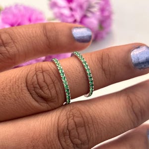 Puede incluir: Dos anillos de plata con una fila de pequeñas piedras preciosas verdes. Los anillos están en un dedo, con un fondo borroso de flores rosas. Las piedras preciosas son de color verde vibrante.