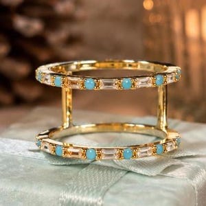 May include: A gold-toned double-band ring featuring two parallel circular bands connected by vertical bars. The bands are set with alternating clear baguette-cut stones and small round turquoise-colored stones arranged in a repeating pattern.