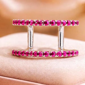 May include: A silver ring with two parallel bands of small, bright pink gemstones. The ring is set on a soft, pink surface, with a blurred floral background.