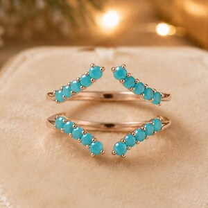 May include: A rose gold ring featuring two chevron-shaped bands, each set with a row of small, round, bright turquoise stones. The ring has an open design, with the two bands meeting at the sides to form a pointed, V-shaped silhouette.