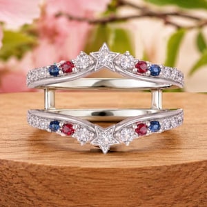 May include: A silver-toned double band ring featuring a V-shaped design. The bands are adorned with clear, round-cut stones, interspersed with small red and blue circular stones. Each band features a central star-shaped setting with a clear stone.
