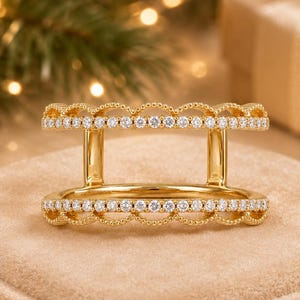 May include: A gold ring guard featuring two parallel bands connected by vertical bars. Each band is adorned with a row of small, clear, round-cut stones and finished with a delicate scalloped edge along the top and bottom of the ring structure.