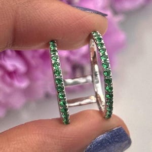 May include: A silver-toned double band ring featuring two parallel rows of small, round, vibrant green stones set along the outer edges of each band, connected by two thin silver-toned metal bars positioned between the two rings.