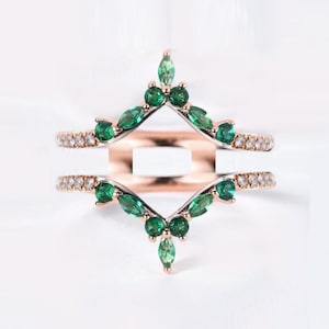 May include: A rose gold ring guard with a double chevron design. The top band features marquise-cut emerald green gemstones and round white gemstones. The bottom band has a similar design. The ring guard is set against a white background.