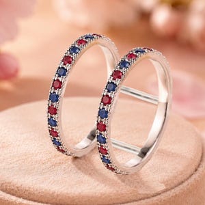 May include: Two thin silver rings featuring an alternating pattern of small, round, faceted red and blue gemstones set along the entire outer band. The rings are positioned upright, one slightly behind the other, showcasing the metallic finish.