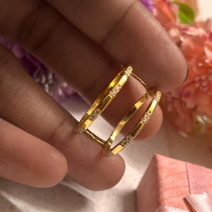 Puede incluir: Un par de pendientes de aro dorados con pequeñas piedras preciosas transparentes engastadas a lo largo de los bordes exteriores. Los pendientes tienen forma rectangular con esquinas redondeadas. El fondo incluye una mano y flores rosas y moradas.