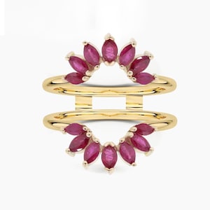 May include: A gold ring enhancer with two bands, each adorned with a half-circle of marquise-cut, deep red gemstones. The gemstones are set closely together, creating a radiant, sunburst-like design. The ring is set against a white background.