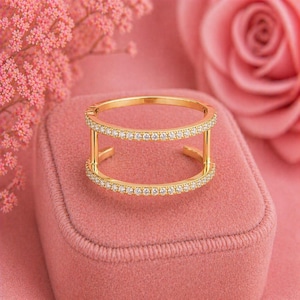 Puede incluir: Un anillo dorado con dos bandas paralelas, cada una incrustada con pequeñas piedras preciosas transparentes. El anillo tiene un diseño abierto y moderno, sobre un fondo rosa con una rosa y elementos florales. Joyería.