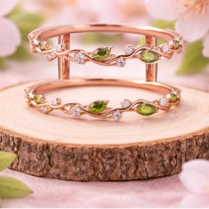 Könnte beinhalten: Ein Ring-Set aus Roségold mit zwei durch vertikale Stege verbundenen Ringen. Die Ringe sind mit einem rankenartigen Motiv gestaltet, das mit kleinen, runden, klaren Steinen und marquise-geschliffenen grünen Steinen in einem zarten Muster verziert ist.