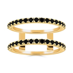 Può includere: Un anello a doppia fascia con una montatura color oro e una fila di piccole pietre nere su ogni fascia. Le due fasce sono collegate da barre verticali dorate, creando un design moderno. L'anello è su sfondo bianco.