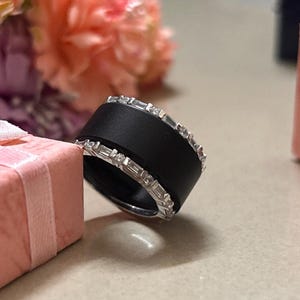 Puede incluir: Un anillo negro ancho con una banda de gemas claras y brillantes. El anillo se encuentra sobre un fondo suave con una caja de regalo rosa y flores. Las gemas parecen ser de talla baguette.