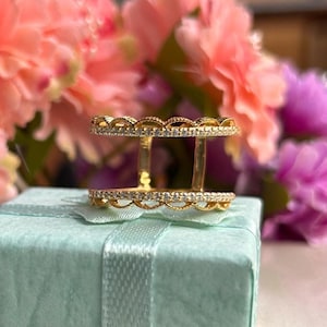 Puede incluir: Un anillo dorado de doble banda con borde festoneado, incrustado con pequeñas piedras preciosas transparentes. El anillo se exhibe sobre una caja azul claro con una cinta, con un fondo borroso de flores rosas y moradas.