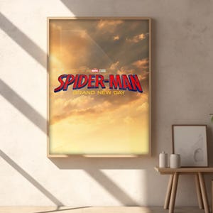 May include: A rectangular framed wall art print featuring a golden sunset sky background with clouds. The text reads Marvel Studios Spider-Man Brand New Day in bold red and blue lettering centered in the middle of the print within a light wood frame.
