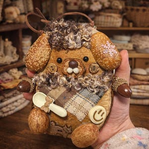 Puede incluir: Adorno artesanal en forma de perro, caprichoso, con un cuerpo marrón texturizado. Presenta pelo rizado marrón, adornos de bellotas y una pequeña cuchara blanca. El perro tiene ojos de botón y un detalle de cara sonriente, con un lazo de cuero marrón.