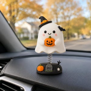 May include: A white ghost figurine with a black witch hat, holding a small orange jack-o'-lantern. The ghost is attached to a metal spring on a black base featuring a small pumpkin, a gray tombstone with the text BOO!, and a white star.
