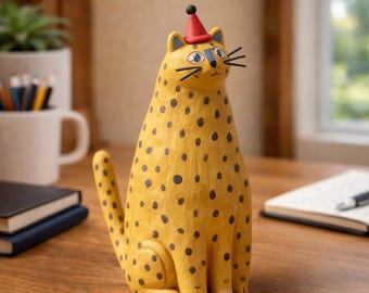 May include: A whimsical, yellow cat figurine with black spots and a red party hat. The handcrafted cat has a cheerful expression with painted whiskers and blue accents. This decorative item is a unique addition to any home decor collection.