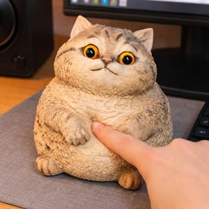 Könnte beinhalten: Detaillierte Figur einer pummeligen Katze mit großen, orangefarbenen Augen und einem leicht mürrischen Gesichtsausdruck. Die Katze ist hellbraun mit dunkleren Streifen und wird von einem Finger berührt. Die Figur steht auf einer grauen Oberfläche.