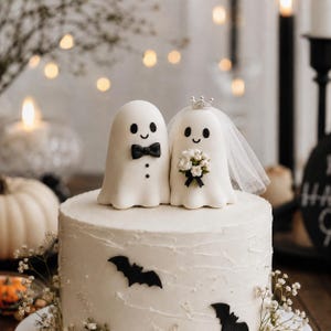 May include: A white cake topped with two ghost figurines. One ghost wears a black bow tie and two black buttons. The other ghost wears a small silver crown, a white tulle veil, and holds a bouquet of white flowers. Black bat cutouts decorate the cake.