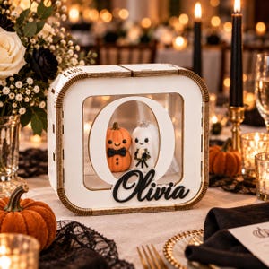 May include: A square white wooden display box featuring a large cutout letter O. Inside the box are two small figures: an orange pumpkin wearing a black bowtie and a white ghost wearing a veil. The black script name Olivia is attached below.