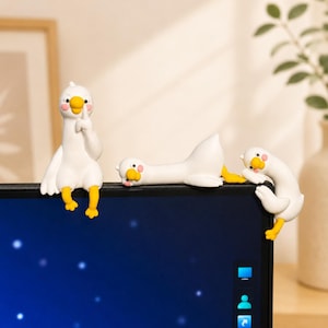 May include: Three white goose figurines with yellow beaks and feet and pink cheeks perched on the top edge of a computer monitor. One goose sits upright with a finger to its beak, one lies flat on its stomach, and one hangs over the side.