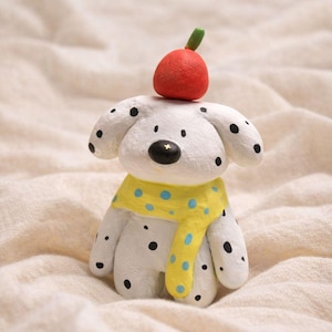 Puede incluir: Figura de perro de cerámica hecha a mano. El perro blanco tiene manchas negras, una bufanda amarilla con lunares azules y una manzana roja en la cabeza. La nariz del perro es negra con una estrella.