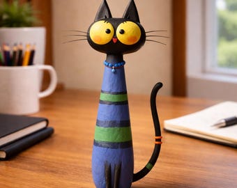 May include: A whimsical black cat figurine with large yellow eyes and a blue and green striped body. The cat has a long, curved tail with an orange accent. It wears a blue beaded necklace. The figurine is on a wooden surface.