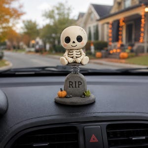 May include: A bobblehead figurine featuring a small, cream-colored skeleton with large black eyes sitting on a metal spring. The base is a gray tombstone with the text RIP, decorated with a small orange pumpkin and a green plant on a round base.