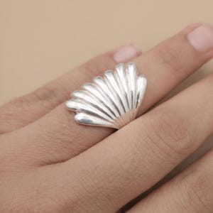 May include: A polished silver ring featuring a fan-shaped design with multiple raised, vertical ridges that resemble the texture of a seashell. The metallic surface is smooth and reflective, covering the width of the finger with a wide, curved band.