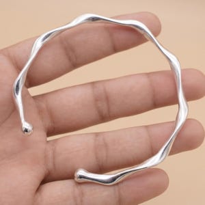 May include: A silver-toned cuff bracelet featuring a continuous, fluid, and wavy design. The metal band has a smooth, polished finish with rounded, bulbous ends at the opening, creating an organic, irregular shape that curves around the wrist.